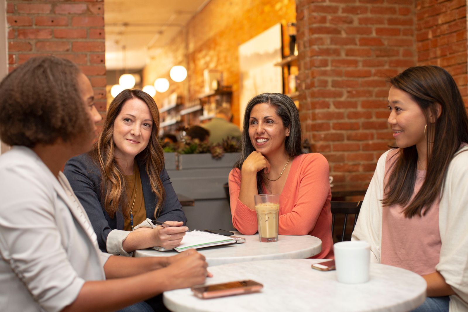 Four women of different races and ages smiling and talking around a cafe table with coffee and a notebook